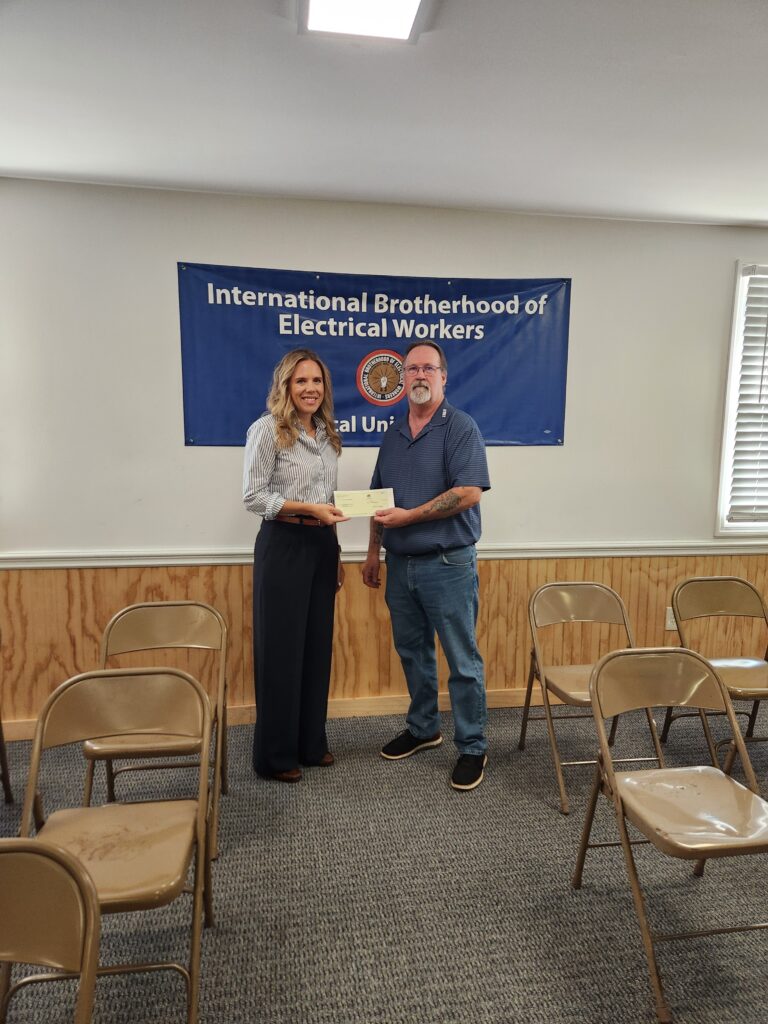 A woman and a man stand in a room and hold a check, with an International Brotherhood of Electrical Workers banner on the wall and folding chairs in the foreground.