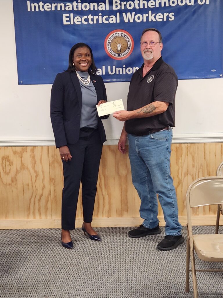 A woman in a dark suit and a man in a black polo and jeans hold a check in front of an International Brotherhood of Electrical Workers banner.